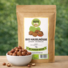 Bio Haselnüsse naturbelassen von Awesome Nuts im Beutel neben einer Schale mit ganzen Haselnüssen auf Holztisch vor grünem Naturhintergrund
